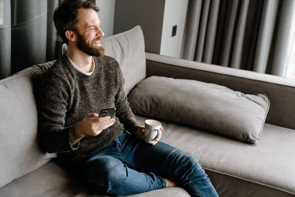 Bearded man using mobile phone and drinking coffee while sitting on sofa at home - Фото, зображення