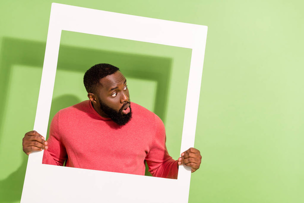 Photo of impressed shocked man hold photo frame look interested empty space isolated on green color background - Foto, imagen