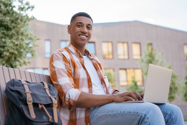 Handsome man copywriter using laptop computer typing, working freelance project looking at camera outdoors. Smiling African American student studying sitting in university campus, education concept - Foto, Imagem