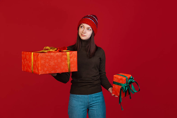Studio shot of cute pretty young girl in warm clothes and knitted hat standing with gift box isolated on red background. - Foto, Bild
