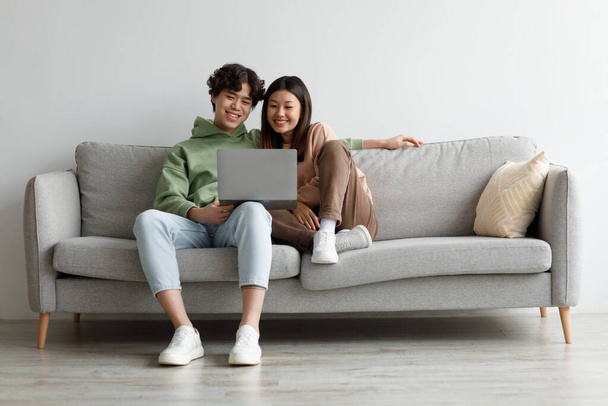 Mutlu genç Asyalı çift dizüstü bilgisayar kullanıyor, evdeki kanepede dinleniyorlar, boşluğu kopyalıyorlar. - Fotoğraf, Görsel