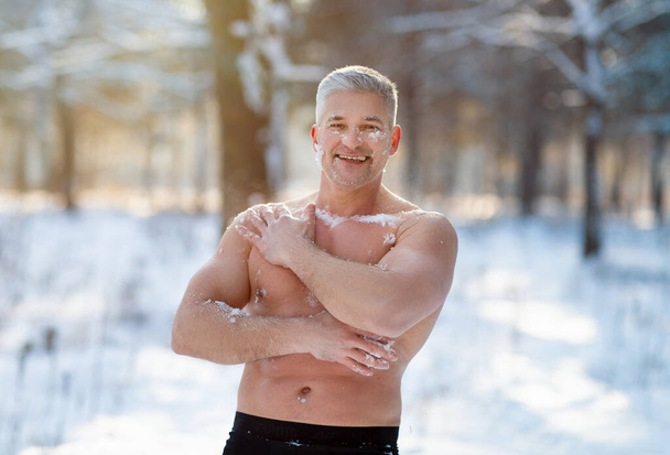 Hombre mayor positivo templando su cuerpo con nieve en el bosque invernal helado. Entrenamiento de exposición en frío - Foto, imagen