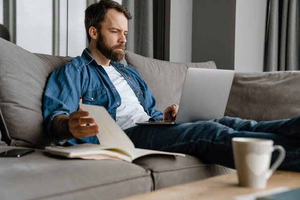 Bearded man writing down notes while working with laptop on sofa at home - Photo, image