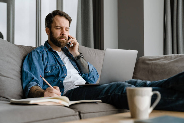 Bearded man talking on cellphone and using laptop while working at home - Foto, immagini