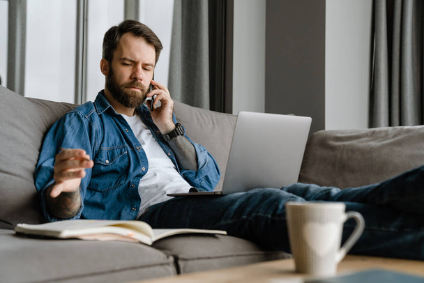 Bearded man talking on cellphone and using laptop while working at home - Foto, Imagem