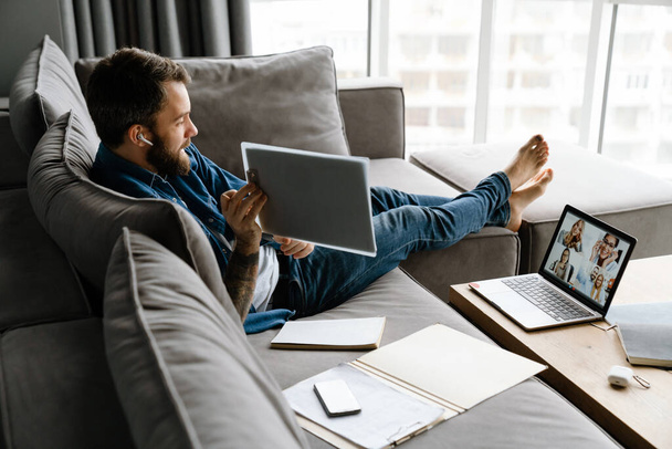 Bearded man making conference call on laptop while sitting on sofa at home - Valokuva, kuva