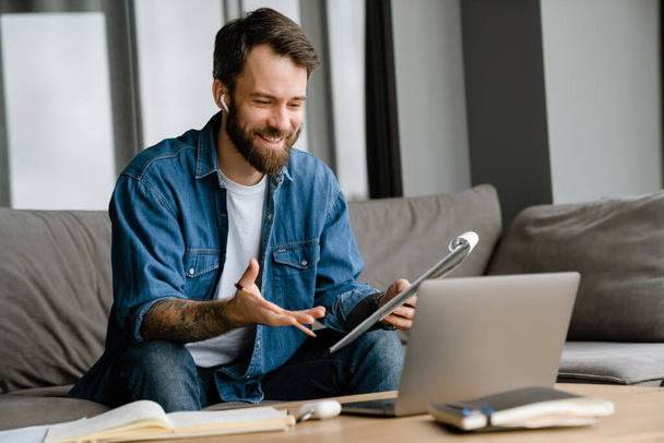 Bearded european man working with laptop while sitting on sofa at home - Photo, image