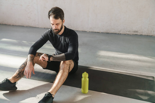 Young brunette bearded sportsman sitting on a fitness mat indoors looking at smartwatch - Foto, immagini