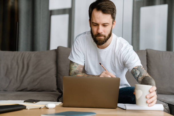 Bearded european man drinking coffee while working with laptop at home - Zdjęcie, obraz