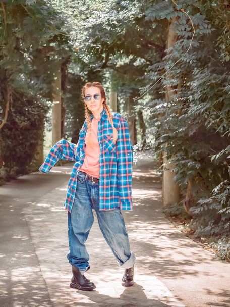 A girl with two pigtails in a plaid shirt with oversleeves and oversized jeans stands in a pose with her legs bent on the concrete path of the park. Street dancer - Fotografie, Obrázek