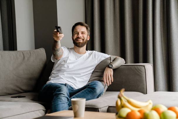 Bearded european man smiling and watching TV while sitting on sofa at home - Fotó, kép