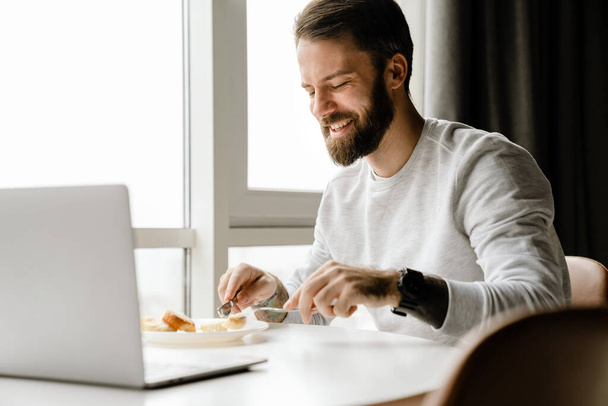 Homme barbu européen travaillant avec un ordinateur portable tout en prenant le petit déjeuner à la maison - Photo, image