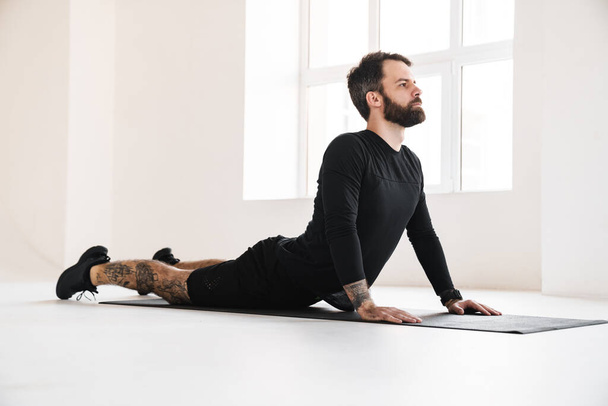 Young brunette bearded man in sportswear stretching while laying on a fitness mat indoors - Foto, Imagem