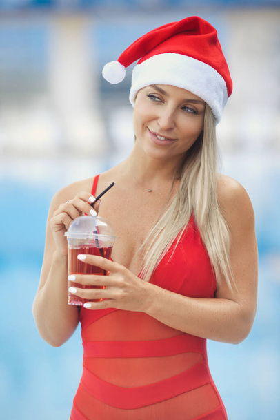 A beautiful blonde woman 25-30 years old is resting in a hotel at the resort by the pool in a red one-piece swimsuit. Happy girl having fun at christmas party with cocktail in santa claus hat. - Fotó, kép