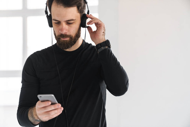 Young brunette bearded white man in sportswear standing indoors listening to music in headphones holding mobile phone - Foto, Imagen