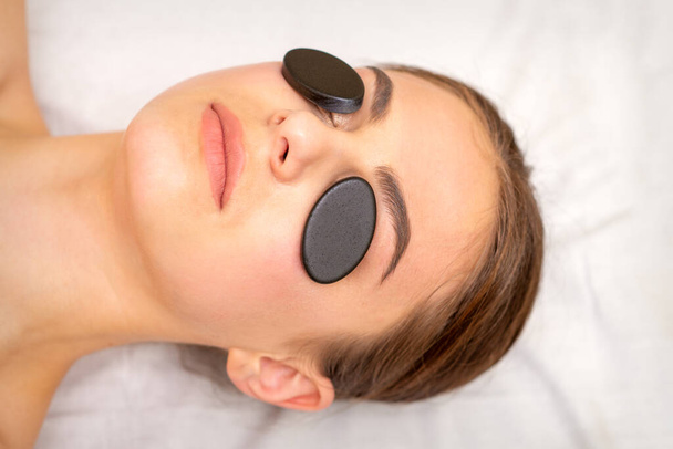 Jeune femme recevant un massage facial avec des pierres minérales noires sur les yeux dans un spa - Photo, image