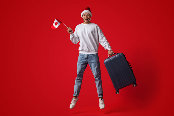 Man draagt Santa Hat springen in de lucht met Canadese vlag en koffer - Foto, afbeelding