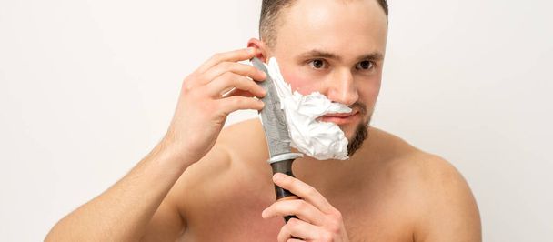 Junger kaukasischer Mann rasiert Bart mit einem großen Messer auf weißem Hintergrund - Foto, Bild