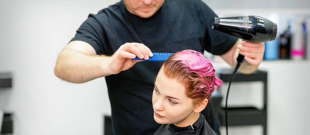 Kampaaja kuivattaa nuoren naisen vaaleanpunaisia hiuksia kauneushoitolassa. - Valokuva, kuva