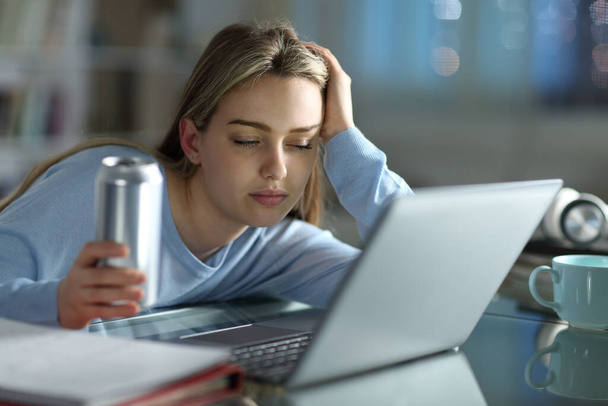 Κοιμώμενος φοιτητής προσπαθεί να μελετήσει με ένα φορητό υπολογιστή κρατώντας ενεργειακό ποτό το βράδυ στο σπίτι - Φωτογραφία, εικόνα