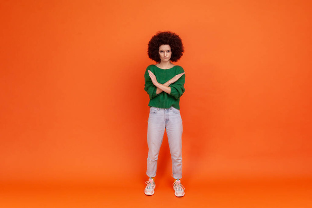 Finish, this is the end. Full length portrait of woman with Afro hairstyle wearing green sweater standing crossing hands, showing x sign meaning stop. Indoor studio shot isolated on orange background. - Photo, image