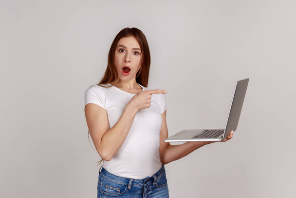 Portrait of amazed emotional woman pointing laptop screen and looking at camera with shocked expression, open mouth, wearing white T-shirt. Indoor studio shot isolated on gray background. - Fotoğraf, Görsel