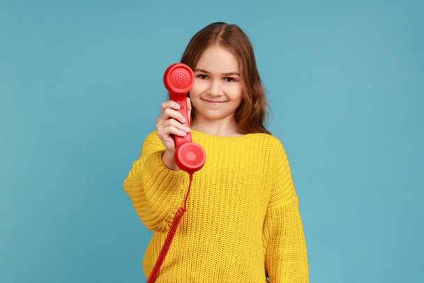 Portrait of little girl holding and showing retro phone handset to camera, asking to answer phone, wearing yellow casual style sweater. Indoor studio shot isolated on blue background. - Photo, Image