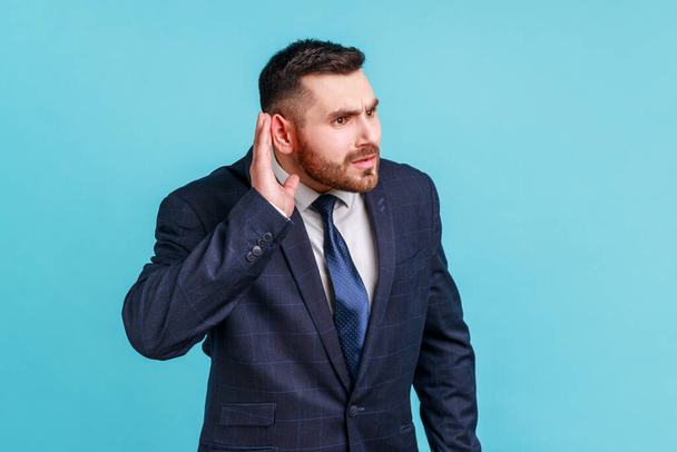 I can't hear you! Portrait of nosy brunette man with beard in suit trying to hear gossip, holding hand near ear and listening attentively carefully. Indoor studio shot isolated on blue background. - Fotó, kép