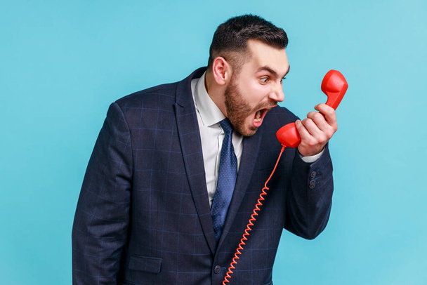 Angry nervous bearded man wearing official style suit screaming and yelling talking retro landline phone, complaining on connection quality. Indoor studio shot isolated on blue background. - 写真・画像