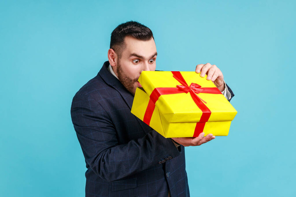 Portrait of curious businessman with present in hands, wearing official style suit, looking inside gift box with shocked expression, unpacking gift. Indoor studio shot isolated on blue background. - Fotoğraf, Görsel