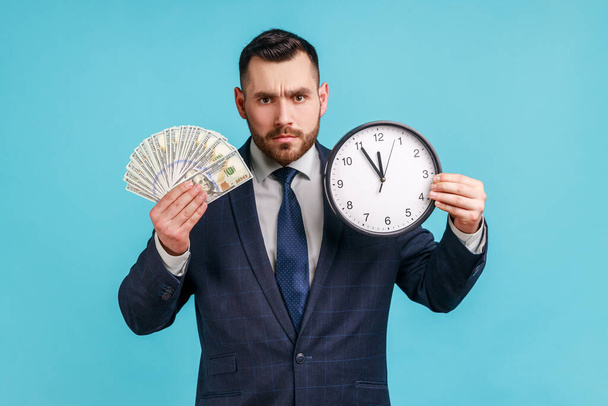 Time to make money! Sad man with beard wearing official style suit holding big clock and dollars banknotes, looking at camera serious facial expression. Indoor studio shot isolated on blue background. - Photo, Image