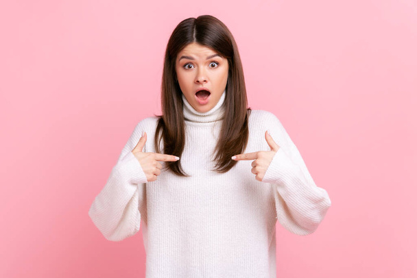 Astonished woman pointing at herself, sincerely surprised by own achievements, shocked expression, wearing white casual style sweater. Indoor studio shot isolated on pink background. - Фото, изображение