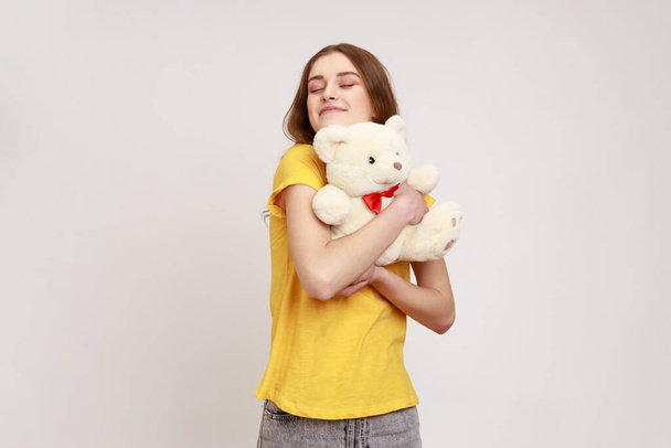 Satisfied happy teenager girl with brown hair in yellow casual T-shirt hugging cute white toy bear, closing eyes and dreaming, enjoying romantic gift. Indoor studio shot isolated on gray background. - Fotoğraf, Görsel