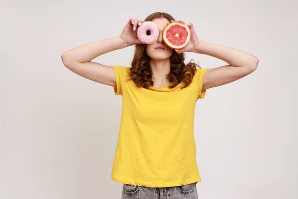 Sugary dessert vs fruits. Portrait of playful brown haired woman in yellow T-shirt covering eyes with fresh grapefruit and sweet doughnut, junk food. Indoor studio shot isolated on gray background. - Zdjęcie, obraz