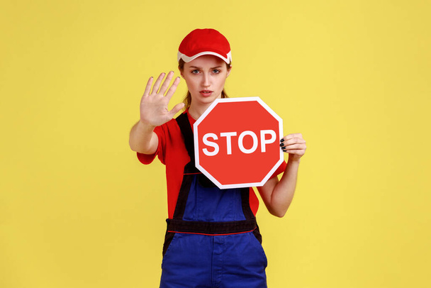 Portrait of worker woman showing stop gesture and holding red stop road sign, prohibitions and restrictions, wearing overalls and red cap. Indoor studio shot isolated on yellow background. - Foto, imagen