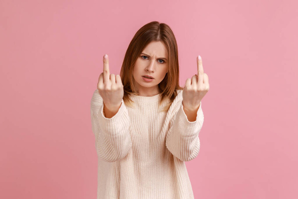 Portrait of blond woman rejecting communication, showing middle finger to express disrespect and hate, vulgar gesture, wearing white sweater. Indoor studio shot isolated on pink background. - Foto, Imagen