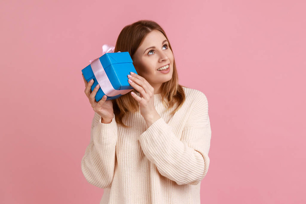 Portrait of attractive curious blond woman shaking wrapped present box, being interested what inside, looking smiling away, wearing white sweater. Indoor studio shot isolated on pink background. - Foto, Imagen