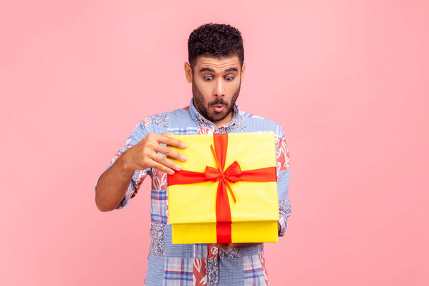 Portrait of curious happy man opening gift, peeking inside box with nosy look, unpacking present, in anticipation of interesting birthday surprise. Indoor studio shot isolated on pink background. - Fotó, kép