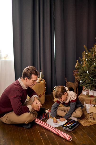 x-mas surprise for beloved people. Father sit with son on floor, child drawing painting. - Photo, Image