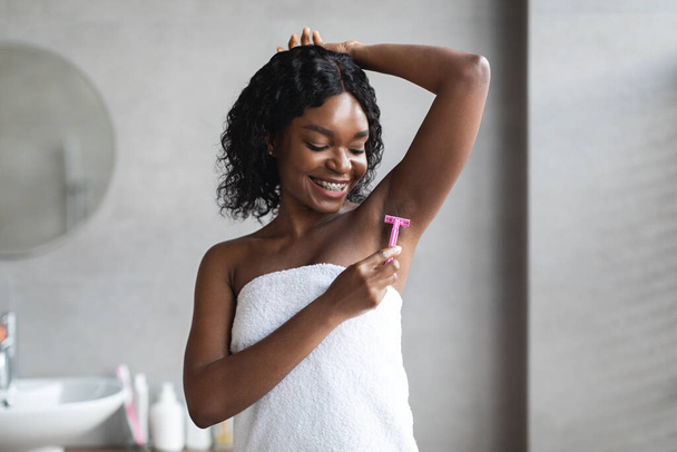 Lächelnde junge schwarze Frau rasiert Achselhöhlen im Badezimmer - Foto, Bild