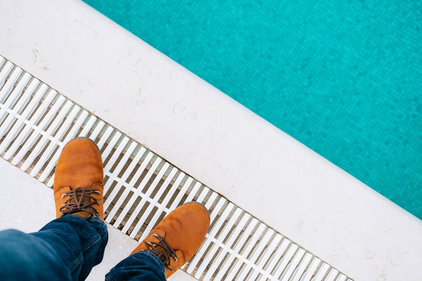 Мужские ноги в коричневых сапогах и синих джинсах стоят на краю бассейна. Крупный план - Фото, изображение