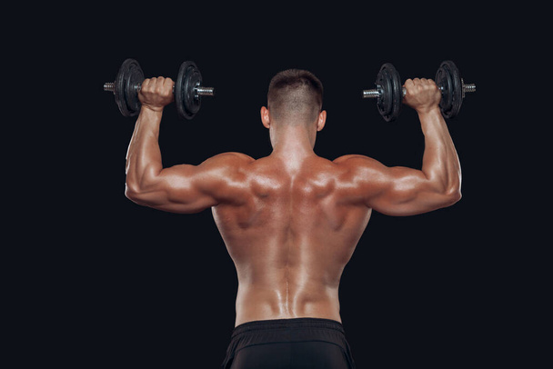 Close up back view of muscular body and strong hands lifting heavy dumbbells isolated over black background - 写真・画像
