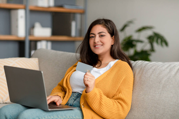 Joyeux arabe femme avec ordinateur portable et achats par carte de crédit en ligne, en utilisant le service bancaire en ligne, assis sur le canapé à la maison - Photo, image