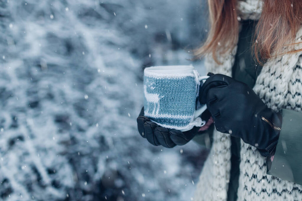 Sluiten van kopje thee of koffie gekleed in gebreide kerstjas in vrouwelijke hand in winterpark. Drankjes om op te warmen in besneeuwd ijskoud weer buiten - Foto, afbeelding