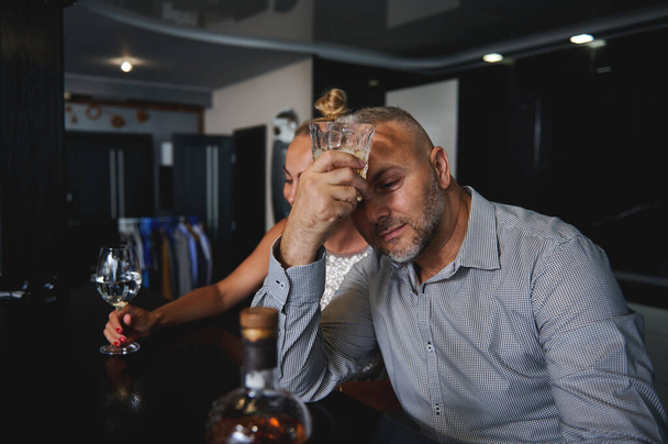 Όμορφος μεσήλικας Ευρωπαίος με περιστασιακό κοστούμι που πίνει αλκοόλ, κάθεται σε ένα πάγκο μπαρ κοντά στην κοπέλα του στο σπίτι - Φωτογραφία, εικόνα