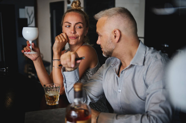 Young blond European woman and handsome Caucasian middle aged man sitting at the bar, talking together, flirting, drinking alcohol, celebrating an event. Heterosexual couple on romantic date - Photo, Image