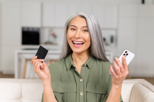 Excited woman holding banking card and smartphone sitting on sofa at home - Foto, Imagem