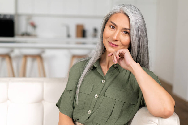 Portrait of attractive middle-aged woman with long silver hair sitting in modern apartment, - Foto, Bild