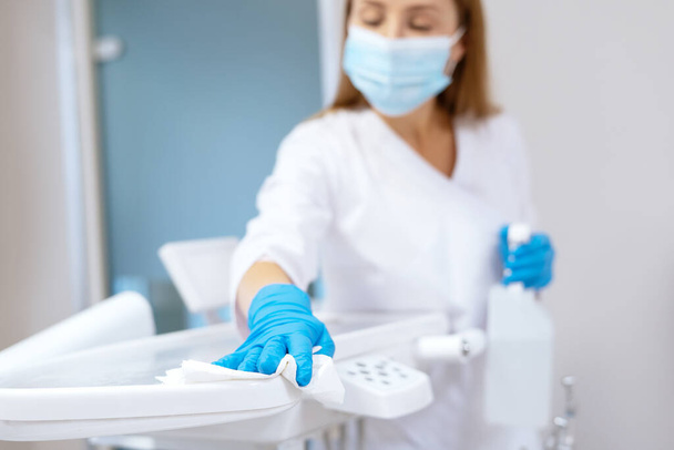 Nurse in protective gloves cleaning work surface at stomatology clinic, sanitizing table with disinfectant spray bottle, washing dental chair with towel, selective focus - Photo, Image