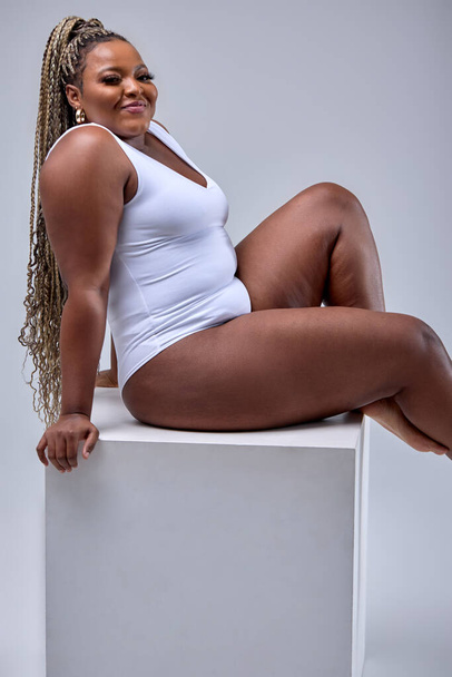 Chubby happy black woman in white bodysuit lingerie posing on white studio background - Fotó, kép